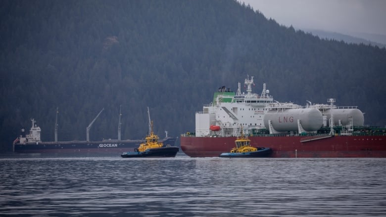 An oil tanker is seen next to two smaller tug boats on a water body, with mountains in the background.