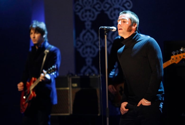 On the right side, a man in focus sings into a microphone during a performance, while on the left, a man in the blurred background plays guitar. 