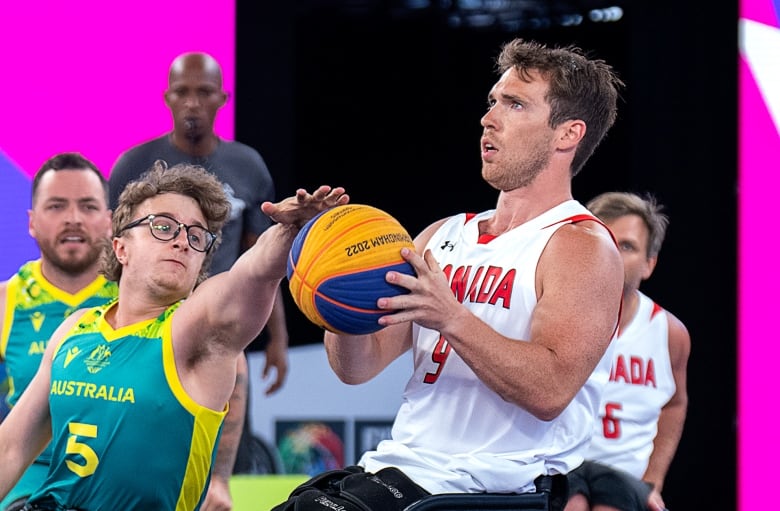 A man in a white Canadian jersey is defended during a basketball game by a man in a green and yellow Australian jersey.