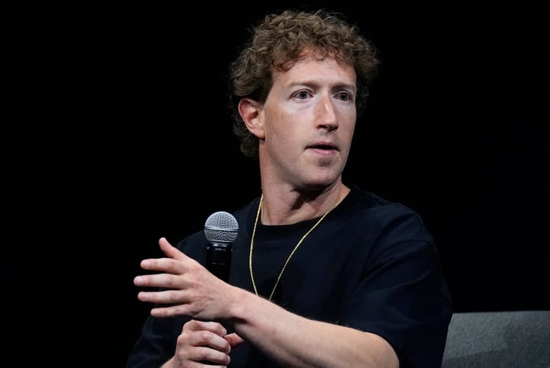 A curly-haired, clean shaven man is shown sitting and gesturing with his left hand while holding a microphone with his right hand.