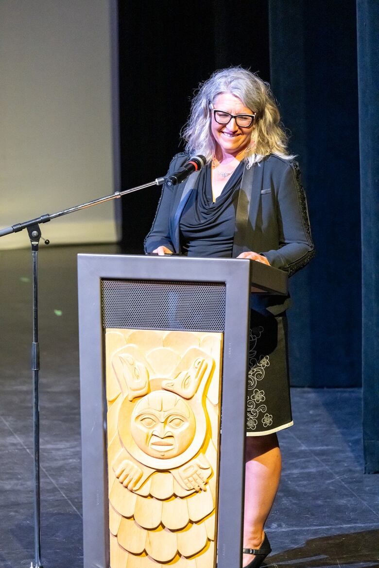 I woman at a podium stands on a stage with a dark background. She wears glasses and a black jacket, she has greying hair.