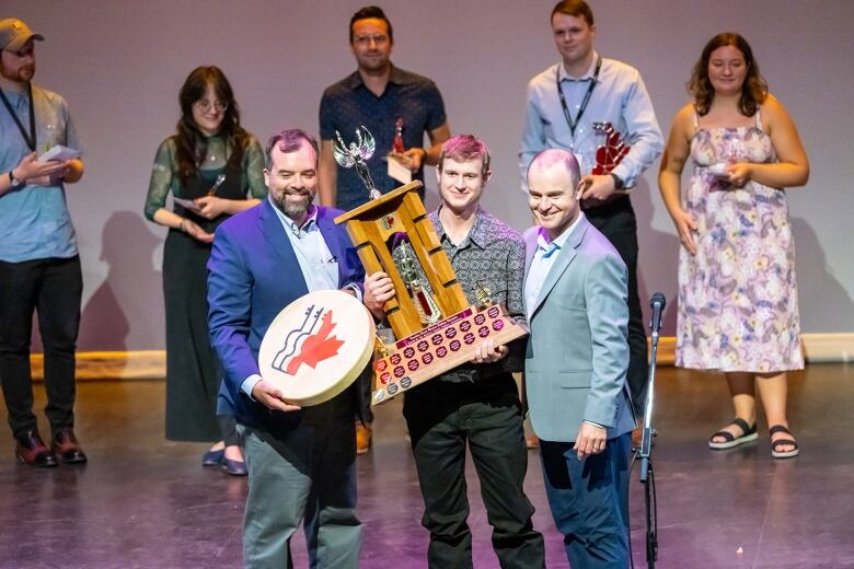 Two men present another with a large award. The award is made of wood with a large figure with wings on the top, there are several labels of past winners on the award.