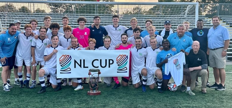 A soccer team poses for a team photo. Players in the front row are holding a sign that reads 'NL CUP' and a trophy sits on the soccer pitch.