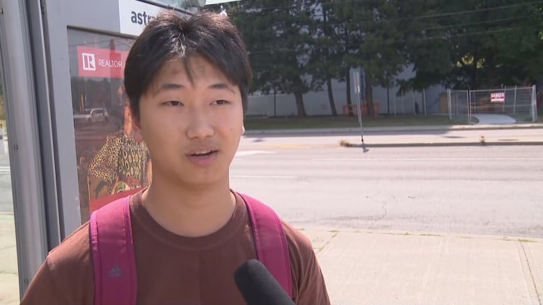 A young man with short black hair stands by a bus stop and speaks into a reporter's microphone