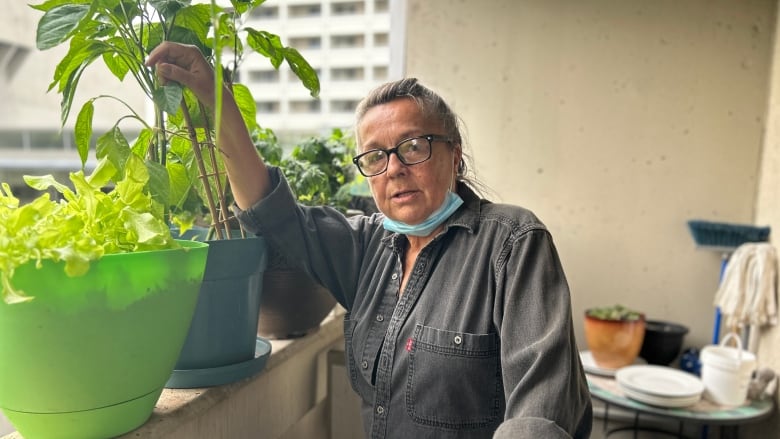 A woman with grey hair and glasses is standing on a balcony with plants.