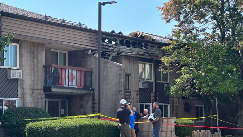 A two-story concrete apartment building with it's roof clearly destroyed by fire. A group of people stand in a circle out front.  The sky is blue and sunny.