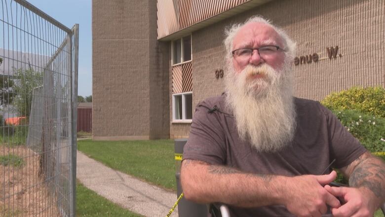 Robert, a middle-aged man with white hair and a long white beard, is seated outside the apartment building next to a fence. 