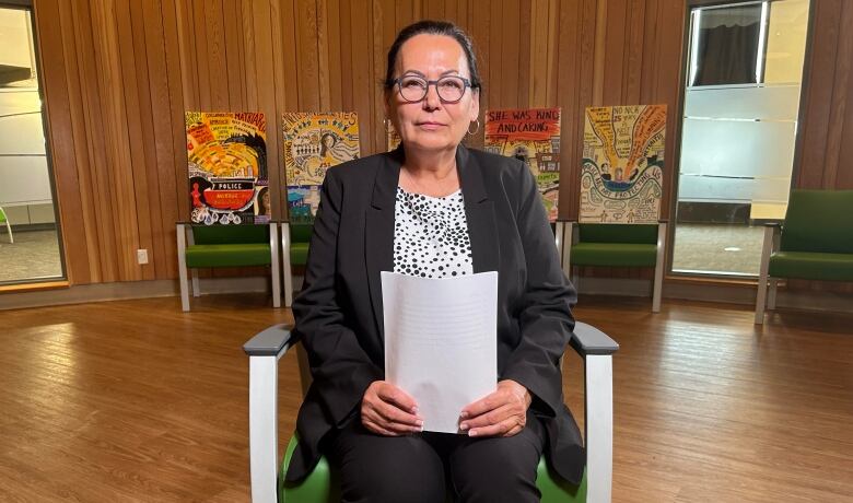 A woman wearing a black blazer and white and black dotted shirt holds papers while sitting in a chair in a room with wood interior.