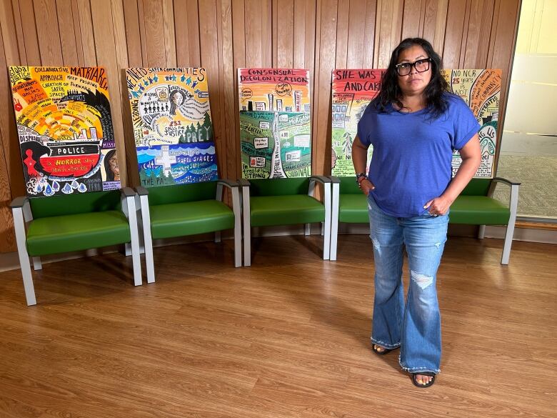 A woman wearing a purple shirt and blue jeans stands in front of five colourful graphic boards placed on chairs.