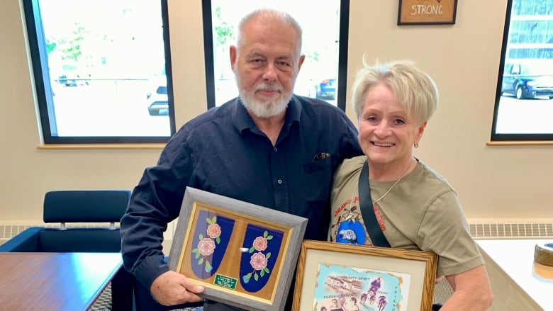 A man and a woman stand in a room holding framed pictures.