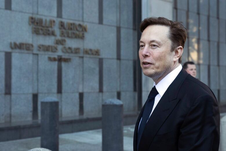 A man in a suit stands in front of a building, which is blurred in the background. 