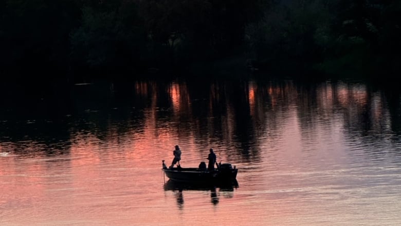 People stand and fish on a boat as the sun sets over the trees behind them.