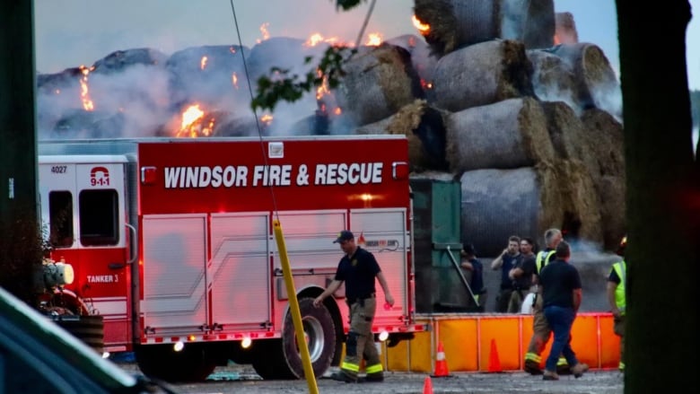 A fire truck backed up against a tall stack of hay bales with fire coming out of the top of the bales.  