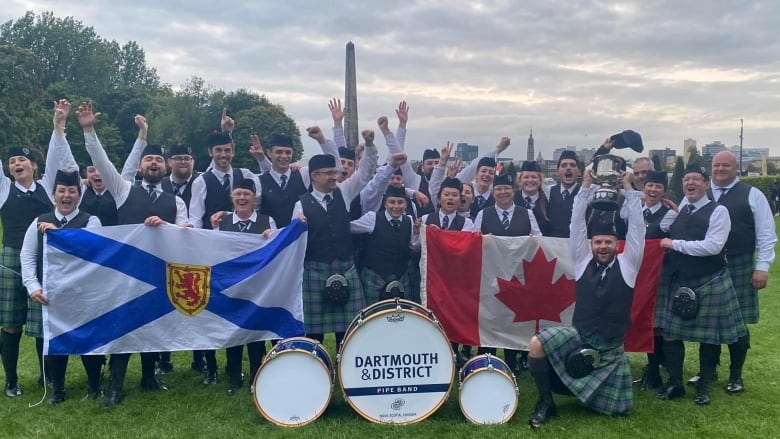 A large group of people wearing formal Celtic garb, including kilts, stand together and cheer. The group holds up the Nova Scotian flag and the Canadian flag.