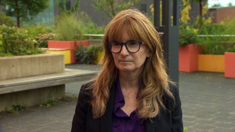 A woman with long red hair is looking off-camera. She is wearing a purple blouse, black jacket, and purple glasses. There are bright orange flower planters in the background.