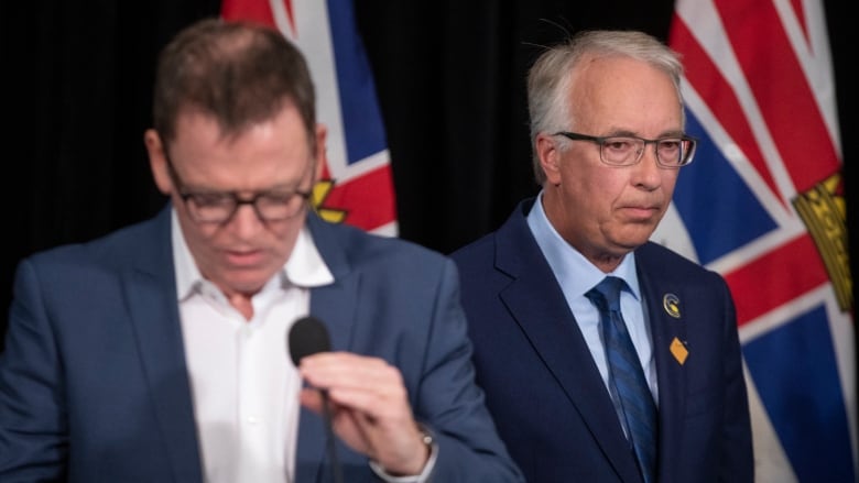 Two men speak with B.C. flags in the background. One of them is in focus and looks pensive, while the other looks down at the microphone.