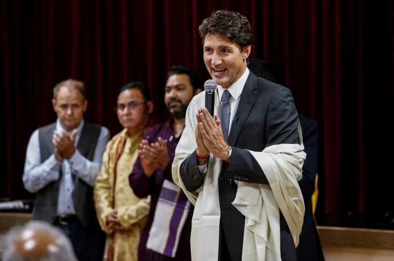 A man in a suit wearing a white shawl around his shoulders holds a microphone in both hands while making a praying gesture.