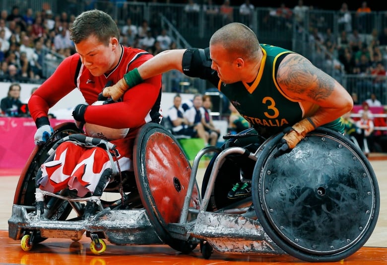 A man wearing red holding a ball in a wheelchair is seen trying to stave off another man in a wheelchair attempting to take the ball away from him.