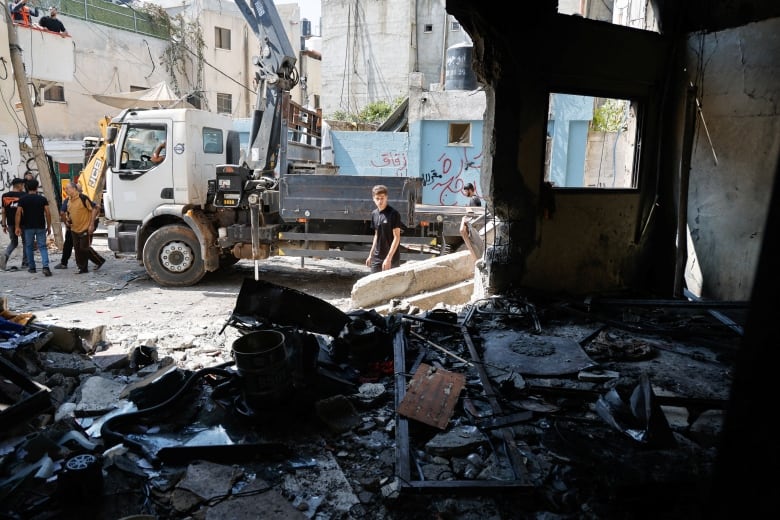 A teenager stands in the middle of rubble, observing damaged buildings around them. Several people are off to the left next to a truck. 