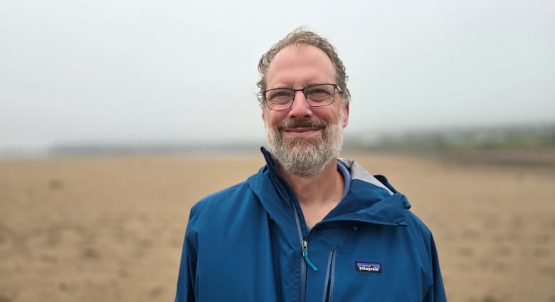 A man with curly brown hair and a greying beard and glasses smiles at the camera while wearing a blue jacket and standing on a beach.