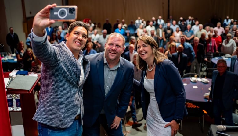 Two males and one female pose for a selfie with a seated crowd behind them.