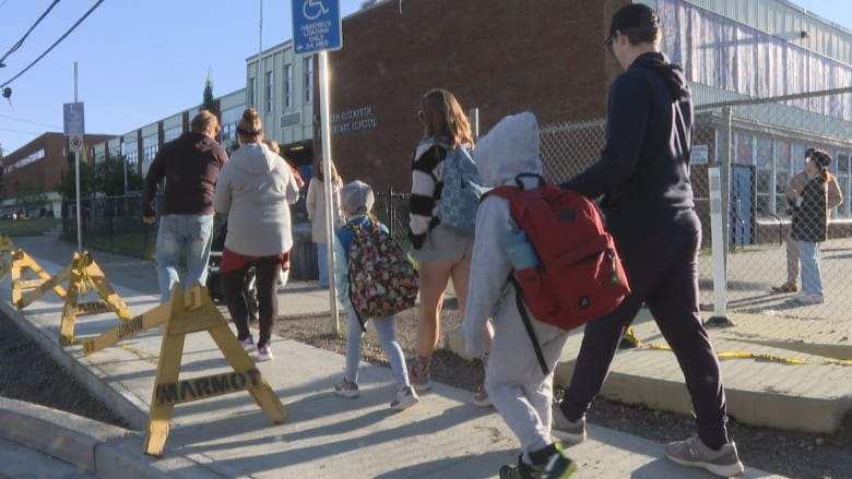 A group of adults and children walk with their backs turned past a fence towards a brick building.