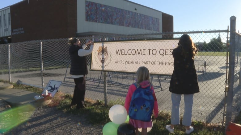 A woman wearing a dark vest over a white shirt and black pants hangs one side of a welcome sign on a chain fence. Another woman wearing a black coat and light-coloured pants hangs the other end. A girl holding multi-coloured balloons wearing a pink jacket and a blue backpack watches.