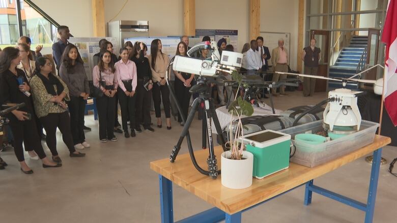 People stand in a room looking at a table full of devices, as well as a plant and soil.