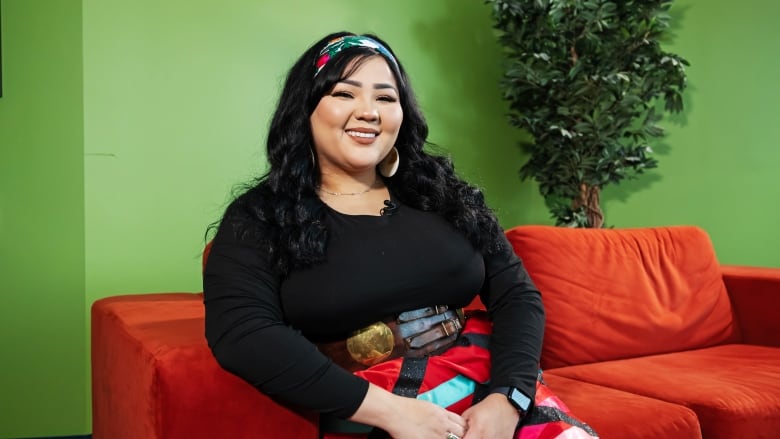 Woman with long, brown hair sits on a red couch. She's wearing a black top and a red, back, blue, white and pink ribbon skirt. 