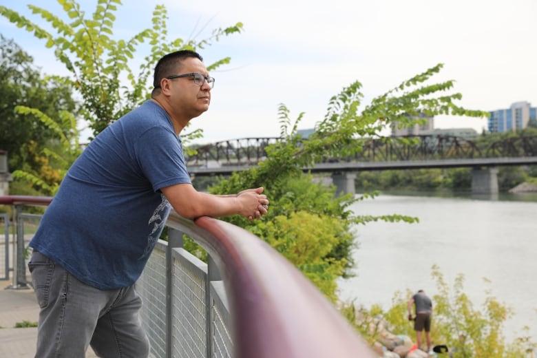 A man in a blue shirt and black glasses leans at a railing by a river, a black bridge stretching out behind him.