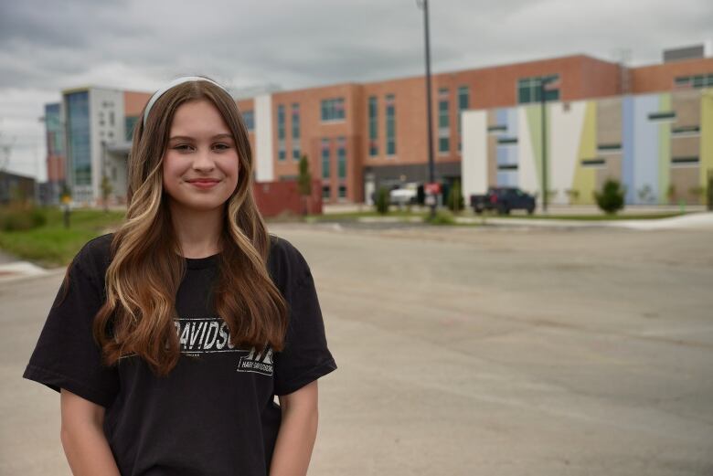 A high school student stands in front of a school that's not quite finished construction. it's a cloudy late summer day.