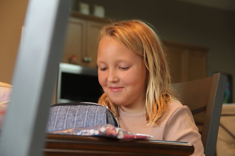 A young girl with blonde hair has a small smile as she looks down at her school supplies laying on the table.