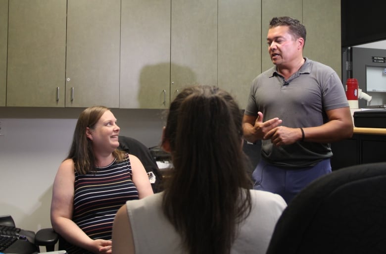 A man in a grey polo is standing in an office, as he speaks to two women who are seated.