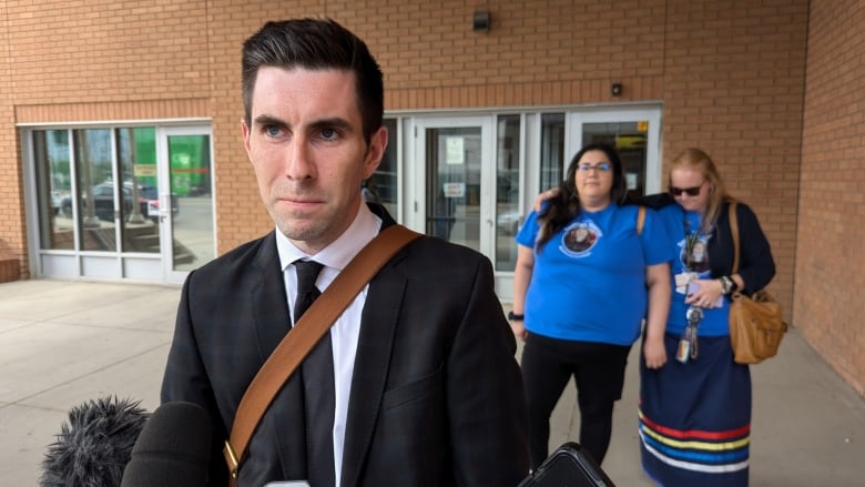 A man in a suit with people standing behind him in blue shirts that read justice for Baeleigh