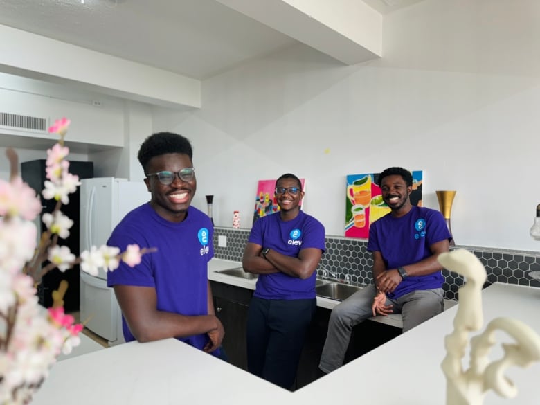 Three men wearing purple shirts stand in a kitchen, smiling with a white counter in front of them.