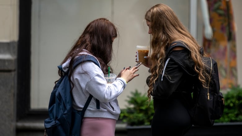 Two young girls are pictured in profile. One of them is holding a cellphone in her hands, while the other holds a cup of iced coffee.