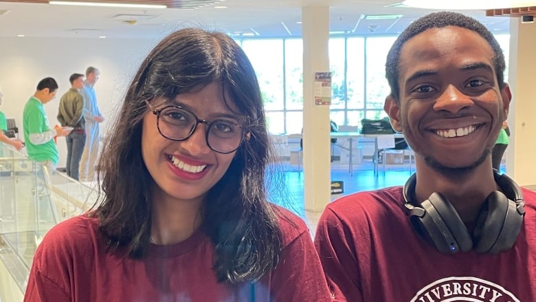 A young woman with long dark hair and glasses stands next to man with headphones around his neck, both wearing burgundy University of Prince Edward Island T-shirts.