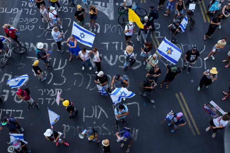 An aerial shot shows people standing on pavement decorated with text. Many are holding Israeli flags. 