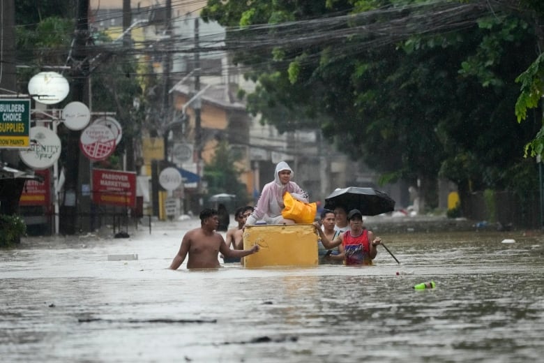 A group of people wade through chest-high water on a flooded street. One person sits on a floating yellow object in the centre of the group. 