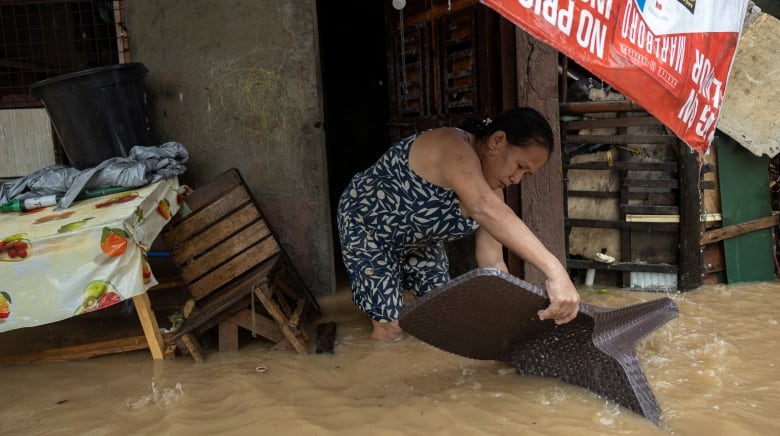 A woman dips a brown chair in calf-high brown water that is flooding the area. 