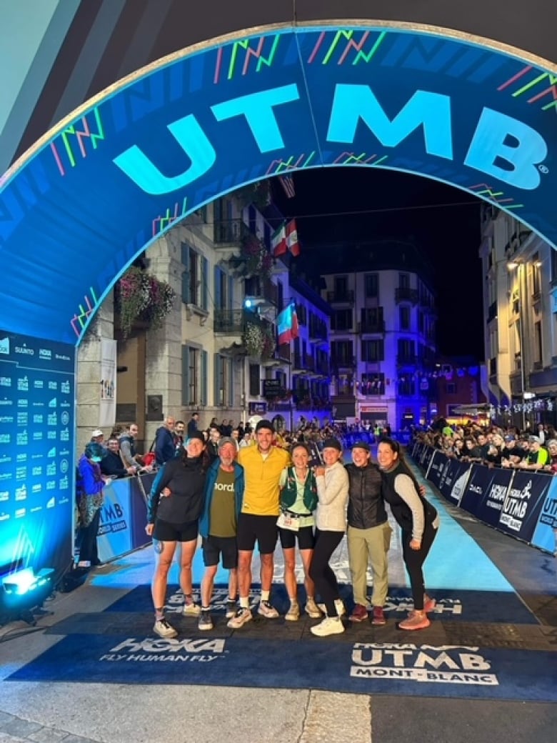 A group of people dressed in athletic clothing stand with their arms around each other underneath a blue banner that says UTMB.