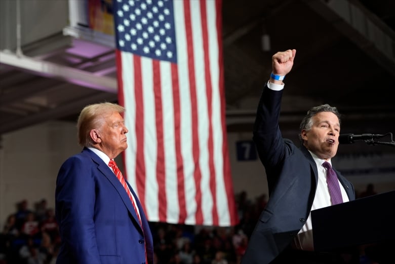 A cleanshaven older man in a suit is shown in profile in front of a massive American flag listening to the side of the stage as another cleanshaven man speaks behind a podium.