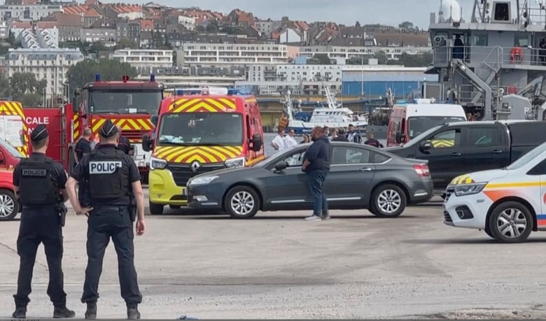 Two police officers stand in the foreground; civilian vehicles and red and yellow emergency vehicles in the background.