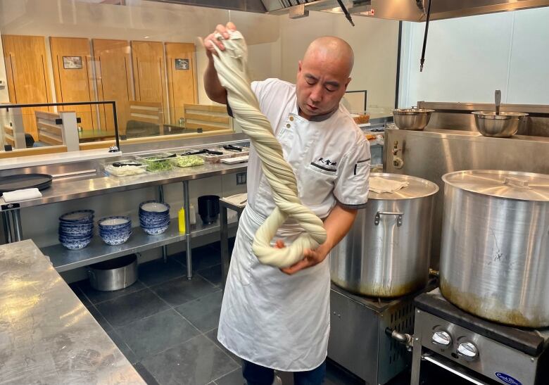 A chef in whites twists around hand-pulled noodles, with vats of food seen behind him.