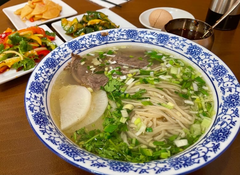 A blue and white ceramic bowl is heaped with soup, noodles and green fresh herbs.