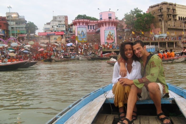 A man and a woman sit on a wooden boat with a busy marketplace behind them.