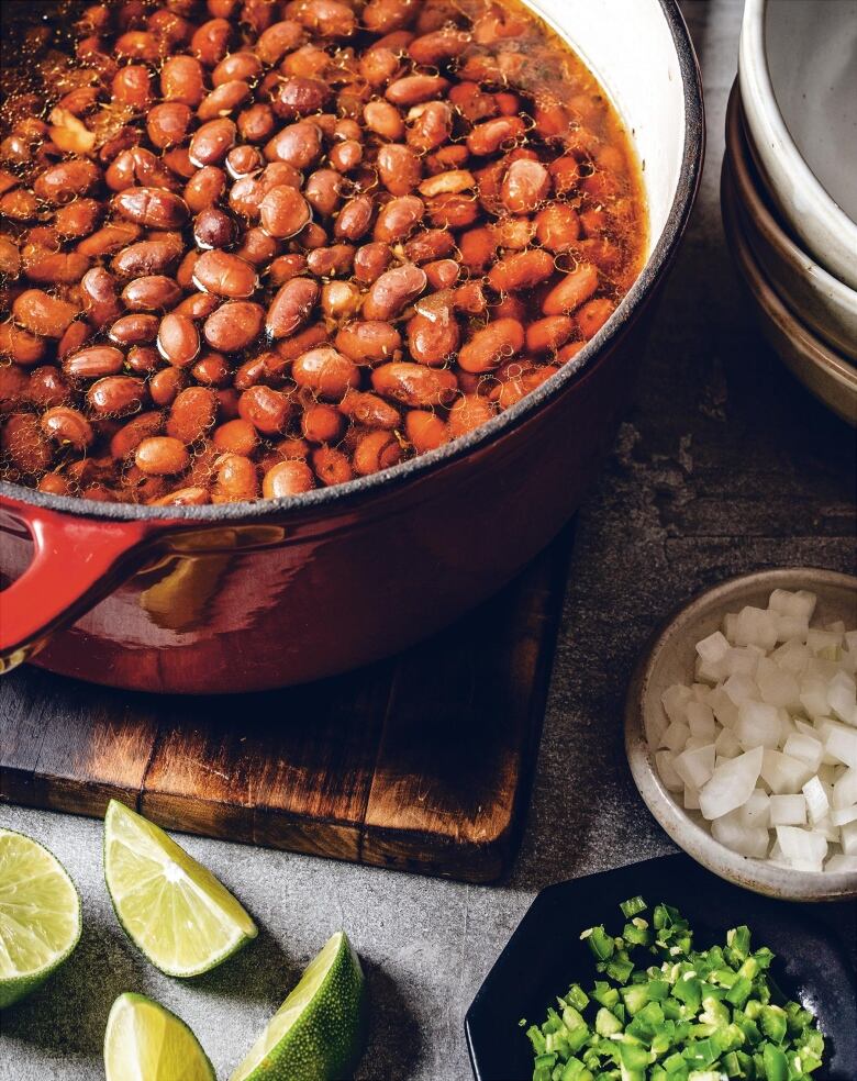 Overhead shot of a red pot of beans on a wooden cutting board next to some lime slices, a stack of bowls and some diced peppers and onions. 