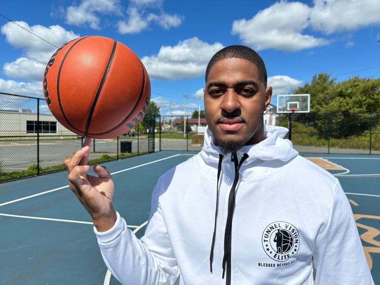 A man is shown in a white hoodie spinning a basketball on the end of his finger.