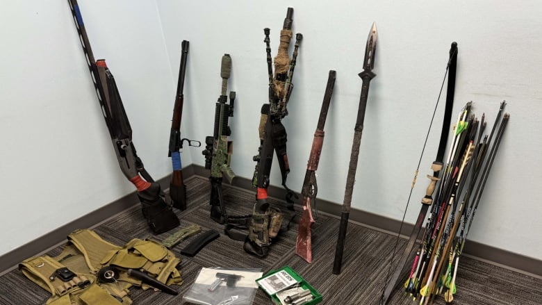 Five long-barreled firearms, a hunting bow, several arrows, a camouflage vest, a handgun, and an assortment of ammunition are displayed against a wall. 