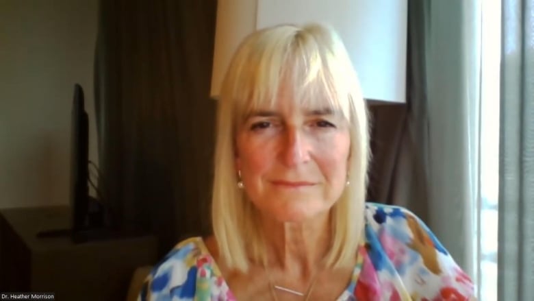 Dr. Morrison sits against the backdrop of a window and lamp wearing a multi-coloured top.